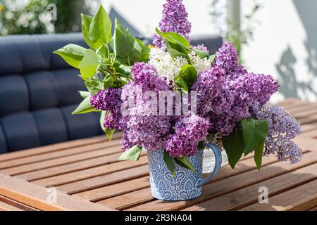 Vista frontale di fiori lilla bouquet in vaso di ceramica su tavolo da giardino in legno contro terrazza offuscata all'aperto nelle giornate di sole Foto Stock