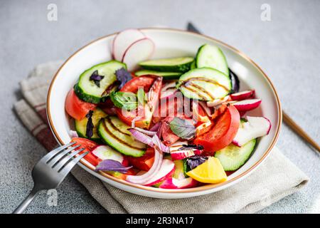 Angolo alto di una gustosa insalata vegetariana con cetriolo, limone, cipolla e pomodoro con foglie verdi e condimento in una ciotola posta sul tavolo Foto Stock