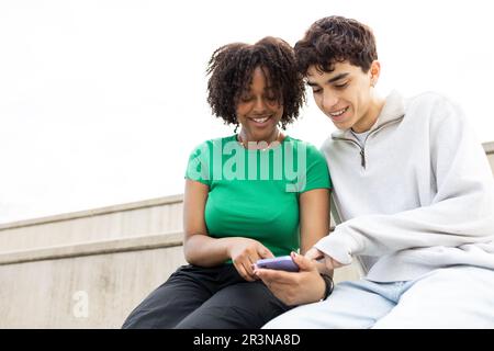 Sorridente coppia multirazziale seduto sulle scale contro il cielo nuvoloso e guardando lo schermo del telefono cellulare mentre si guarda un interessante film insieme dentro Foto Stock