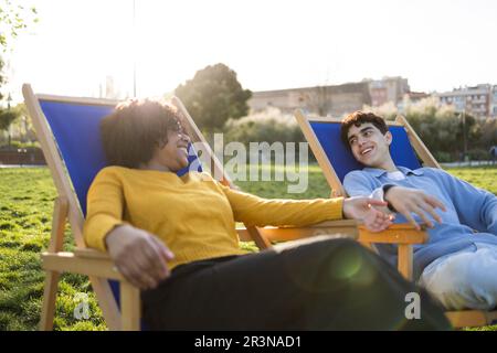 Allegri giovani amici multirazziali maschi e femmine che si rilassano su sedie a sdraio sul campo verde mentre si guardano e si parlano contro città sfocate Foto Stock