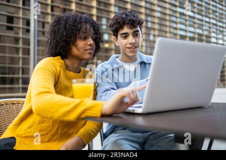 Giovani amici studenti multirazziali seduti al tavolo con bicchiere di succo d'arancia e guardando lo schermo del computer portatile in luce del giorno, mentre la femmina nera punta pinna Foto Stock
