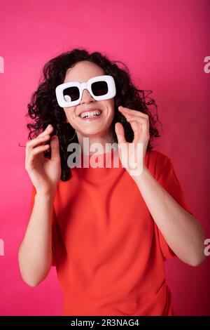 Ritratto di giovane donna eccitata in abito rosso alla moda e grandi occhiali da sole bianchi sorridendo e guardando la macchina fotografica mentre in piedi contro sfondo rosa dentro Foto Stock