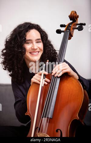 Calma sorridente musicista femminile in abiti neri formali seduti sul divano e suonando violoncello acustico con fiddlestick mentre si guarda la macchina fotografica Foto Stock