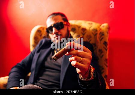 Serio giovane modello maschile bearded in tuta e occhiali da sole seduti in poltrona con sigaro e guardando la macchina fotografica su sfondo rosso Foto Stock