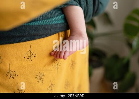 Madre anonima che indossa un supporto avvolgente con una piccola gamba del neonato che si stende fuori mentre si è in piedi in una stanza sfocata Foto Stock