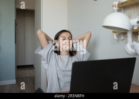 Ragazza adolescente positiva in abiti casual con gli occhi chiusi e le mani dietro la testa contemplando gli studi mentre si siede a tavolo con il computer portatile e il lavoro Foto Stock