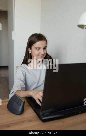 Ragazza adolescente si è concentrata in abbigliamento casual studiando mentre si siede alla scrivania con un notebook e lavorando a un progetto remoto a casa Foto Stock