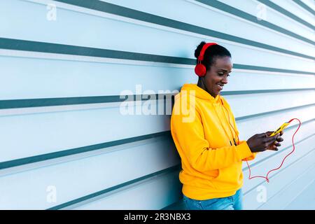 Giovane donna positiva in cuffie sorridente e videochiama con lo smartphone mentre si trova in strada contro la parete blu Foto Stock