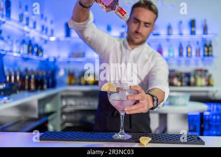 Barkeeper giovane maschio concentrato versare bevande alcoliche dalla bottiglia in un bicchiere di cocktail con una fetta di arancia sul banco mentre si guarda verso il basso contro sfocato Foto Stock