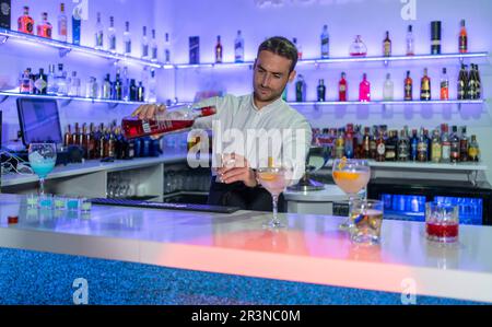 Barista giovane maschio concentrato guardando verso il basso e versando l'alcool dalla bottiglia nel jigger mentre preparano le bevande del cocktail e si levano in piedi in piedi nel banco Foto Stock