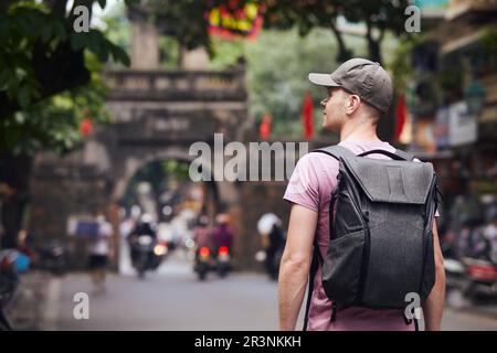Il viaggiatore cammina sulla trafficata strada asiatica. Vista posteriore di uomo con zaino nel centro storico di Hanoi, Vietnam. Foto Stock