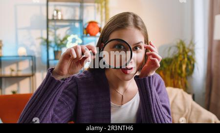 Ricercatore ricercatore scienziato giovane donna con lente di ingrandimento vicino al viso, guardando in macchina fotografica con grande zoomato divertente occhio, la ricerca, l'analisi a casa. Ragazza seduta sul divano nella camera dell'appartamento Foto Stock