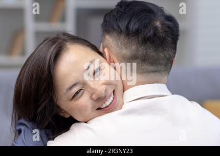 Primo piano. Giovane coppia asiatica, famiglia che abbraccia insieme. Il volto felice di una giovane donna che abbraccia e sostiene il marito, si incontra sorridendo a casa. Foto Stock
