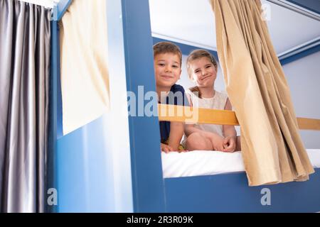 I piccoli caucasici si siedono sul letto a castello e guardano la macchina fotografica con un grande sorriso Foto Stock
