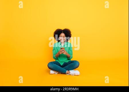 Foto a tutta lunghezza di una donna allegra, trendy, afro-americana o brasiliana, si siede su uno sfondo giallo, utilizza il suo smartphone, parla sui social network, navigando su internet, notizie, sorrisi Foto Stock