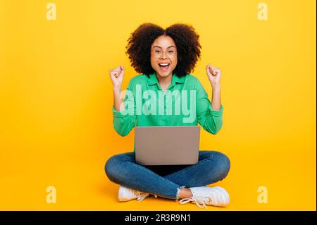 Foto a tutta lunghezza di una gioiosa donna ricciola afro-americana o brasiliana, elegantemente vestita, si siede su uno sfondo giallo con un notebook, si rallegra del successo, gesticula con le mani, sorride alla fotocamera Foto Stock