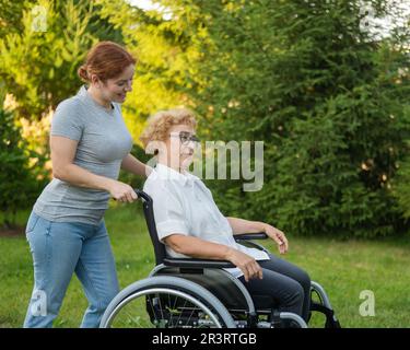 Una donna caucasica trasporta una madre anziana seduta su una sedia a rotelle attraverso il parco. Passeggiate all'aperto. Foto Stock