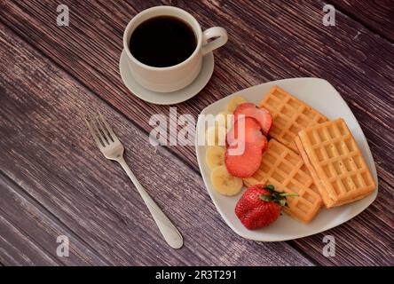 Un piatto con waffle viennesi freschi con fragole e banane su un tavolo di legno, accanto a una tazza di caffè nero caldo su un piattino e una forchetta. Top vie Foto Stock