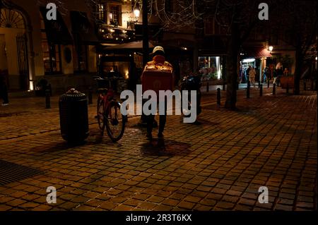 Corriere per un servizio di consegna di cibo in attesa di fronte ad un ristorante di notte a Bruxelles. Addetto alla consegna in uniforme. Foto Stock