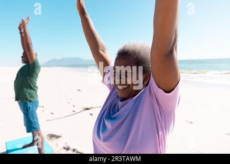Donna e uomo anziano afroamericano sorridente che alzano le braccia e si esercitano in spiaggia contro il cielo limpido Foto Stock