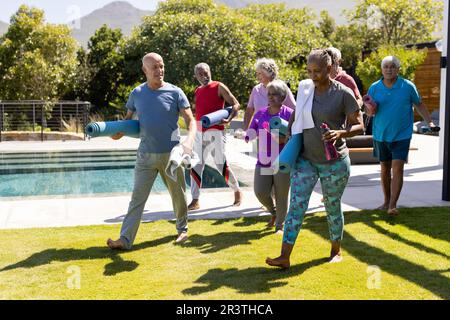 Felice diversi amici anziani in possesso di tappetini yoga, a piedi e parlare dopo sessione in giardino soleggiato Foto Stock