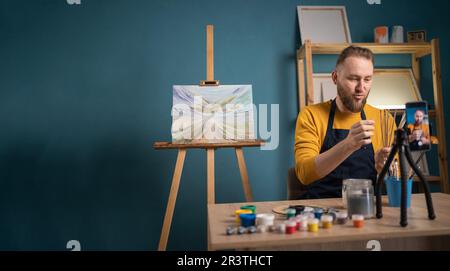 Giovane artista seduto alla scrivania e in studio d'arte per registrare lezioni d'arte online. Blogger fare un video per il suo blog sull'arte che mostra la vernice acquerello Foto Stock