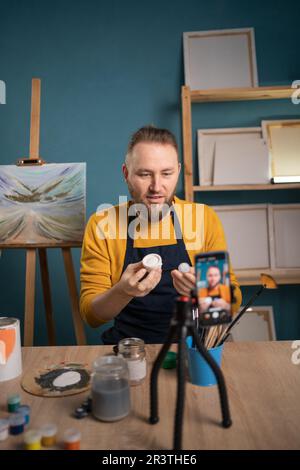 Giovane artista seduto alla scrivania e in studio d'arte per registrare lezioni d'arte online. Blogger fare un video per il suo blog sull'arte che mostra la vernice acquerello. Foto Stock