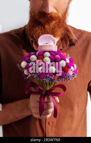 Proposta di matrimonio. Bell'uomo elegante in camicia marrone dona anello d'oro bianco in scatola rosa bel bouquet di fiori. Barbuto ragazzo che propone t ragazza Foto Stock