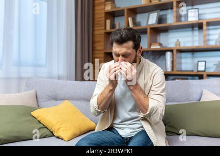 Malato uomo maturo seduto sul divano a casa, l'uomo ha allergia e naso che cola, starnuti in salotto da solo. Foto Stock