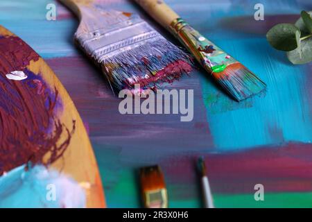 Tela con pittura astratta colorata, diversi pennelli e tavolozza di artisti in legno, primo piano Foto Stock