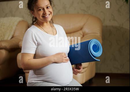 Felice donna incinta sorride guardando la macchina fotografica, tiene un tappetino fitness e le carezza delicatamente la pancia, godendo la sua gravidanza Foto Stock