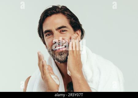 Felice uomo caucasico è orgoglioso della sua routine di cura, applicando con cura la crema per la barba. Con una mano ferma e uno sguardo focalizzato, si preparò Foto Stock