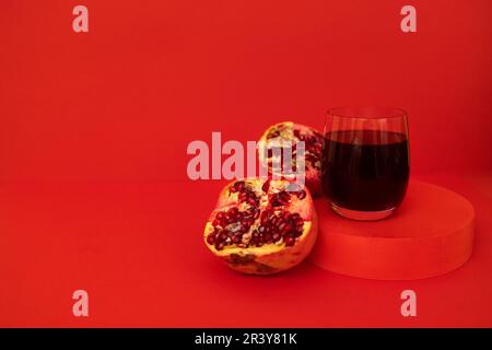 Un bicchiere di succo di melograno su un podio con melograno diviso in pezzi. Sfondo rosso luminoso. Foto Stock