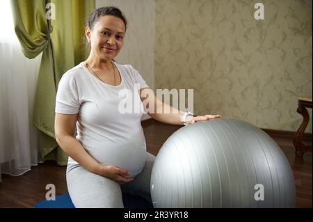 Autentica donna incinta sorride alla macchina fotografica, tocca il ventre, seduto su un tappetino yoga mentre si esercita con la palla fit Foto Stock