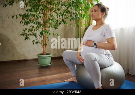 Donna in gravidanza attiva tiene le mani sul ventre, esegue esercizi di respirazione prenatale, stretching e rilassamento su una palla Foto Stock