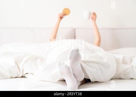 Mano femmina che tiene una tazza di caffè e cracker da sotto una coperta nel letto. Donna che si sveglia al mattino Foto Stock