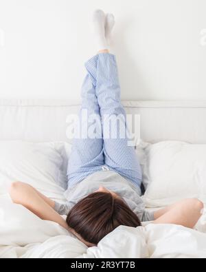 Le gambe femminili si alzarono in alto e le braccia sotto la testa sdraiati sul letto in camera da letto indossando pigiami Foto Stock