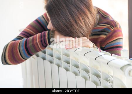 Donna che indossa un pullover seduto vicino al radiatore del riscaldatore e lo abbraccia Foto Stock