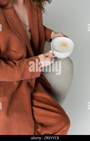 vista ritagliata della giovane donna in terracotta e abito alla moda con collana che tiene il bicchiere di cappuccino con schiuma di latte mentre si siede in comoda poltrona Foto Stock