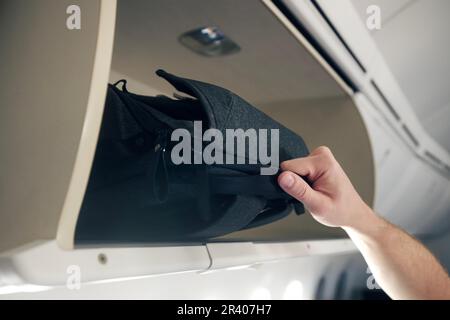 L'uomo viaggia in aereo. La mano più vicina del passeggero sta mettendo il bagaglio a mano negli armadietti sopra i sedili dell'aereo. Foto Stock