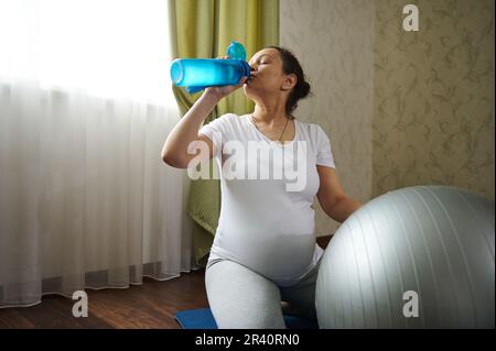 Multi-etnico donna incinta beve acqua dalla bottiglia, seduta sul tappeto blu, rinnovando l'equilibrio dell'acqua dopo la gravidanza fitness Foto Stock