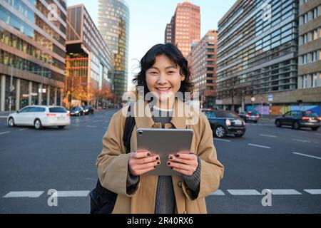 Ritratto della studentessa asiatica, si trova nel centro della città con le auto sulla strada trafficata, tiene il tablet digitale e sorride alla fotocamera Foto Stock