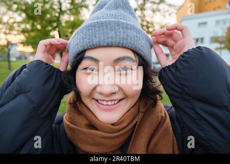 Ritratto di carina ragazza asiatica in cappello e sciarpa, passeggiate in città in clima di primavera freddo, sorrisi e sembra felice, si trova nel parco n Foto Stock