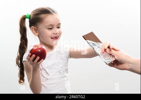 Ritratto di una bambina su sfondo bianco, sceglie il cioccolato su una mela, cibo sano vs cibo spazzatura. Foto Stock