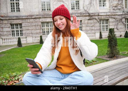 Giovane ragazza sorridente rossa siede sulla panca e utilizza l'app per smartphone, dice ciao e ondate alla fotocamera del telefono cellulare, chat video Foto Stock