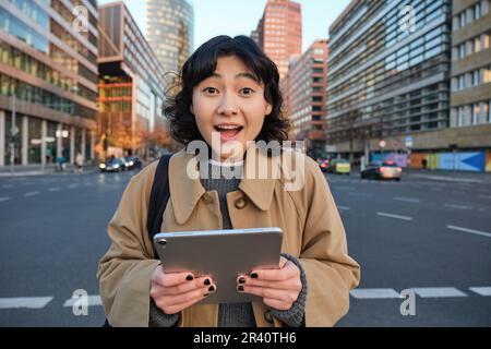 Il ritratto della ragazza coreana sembra sorpreso, ha scoperto qualcosa di straordinario, tenendo il tablet digitale con il volto gioioso, si trova sulla strada Foto Stock
