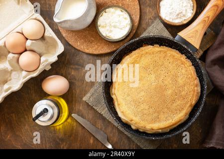 Festeggiamo la giornata delle fritture, cucinando una sana colazione. Deliziose crepes o frittelle fatte in casa in una padella e ingredienti su un r Foto Stock