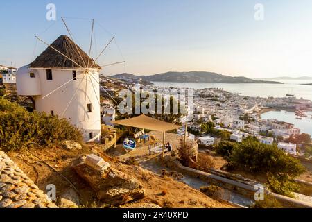 Vista panoramica sulla collina del porto della città di Mykonos dal pittoresco castello veneziano con un mulino a vento in primo piano quando il sole inizia a tramontare. Foto Stock
