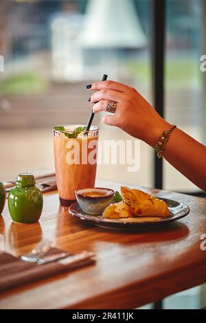 La mano della donna tiene il vetro con il milkshake o il frullato sul tavolo di legno nel caffè sullo sfondo della finestra Foto Stock