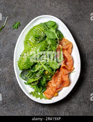 Bassa di un glucide insalata. Gli spinaci, rucola con insalata di avocado e salmone. Nero lo sfondo di calcestruzzo, piastra bianca, vista dall'alto Foto Stock